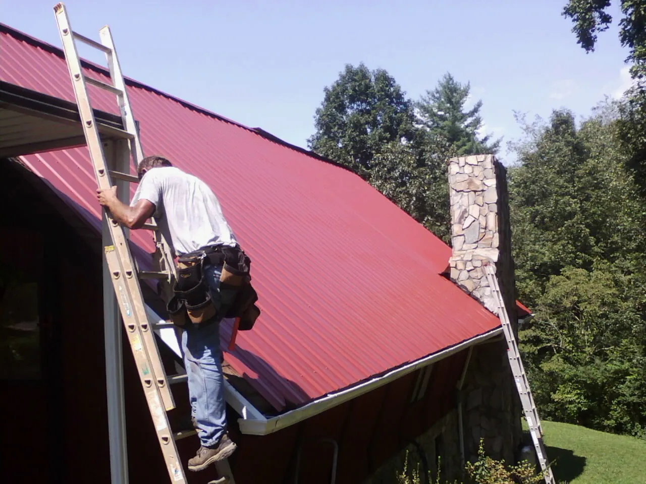 Roofer on a ladder beside a red metal roof and stone chimney — Soffit & Fascia Repair by Roofing Brothers in Buena Vista